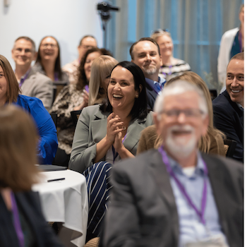 A smiling audience applauding during an Amplify Alliance conference session.